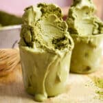 Vegan matcha ice cream overflowing in glass cups, rich and creamy with a vibrant green hue, melting over a wooden board with bamboo whisk and matcha powder in the background.