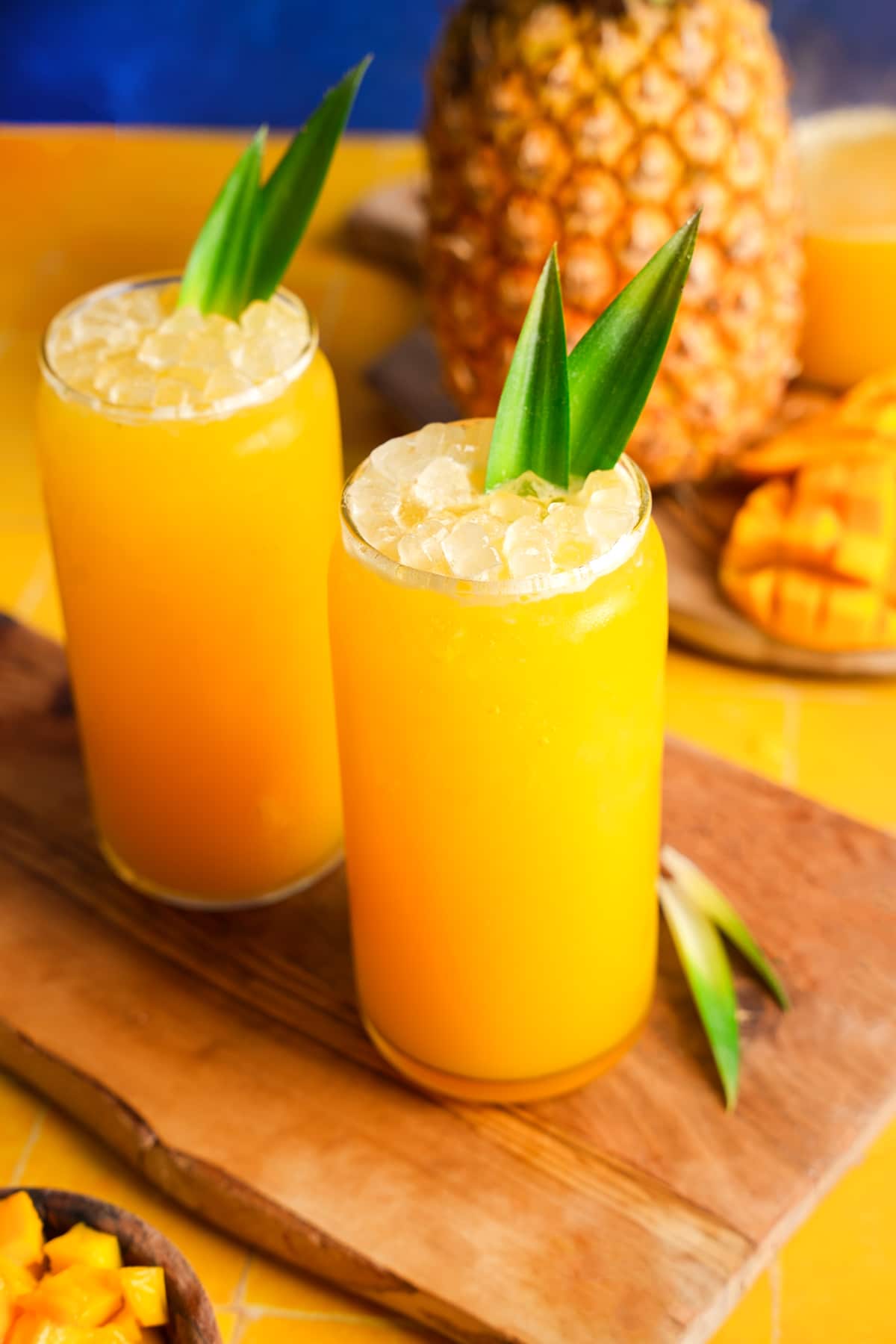 Two tall glasses of fresh pineapple mango juice topped with crushed ice and garnished with pineapple leaves, surrounded by chunks of mango and a whole pineapple in the background on a vibrant yellow surface.