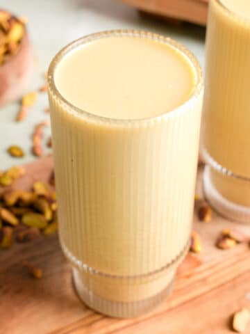Tall glass of creamy pistachio nut milk on a wooden board, surrounded by chopped pistachios and a rustic bowl in the background, showcasing a smooth, rich texture and natural green hue.