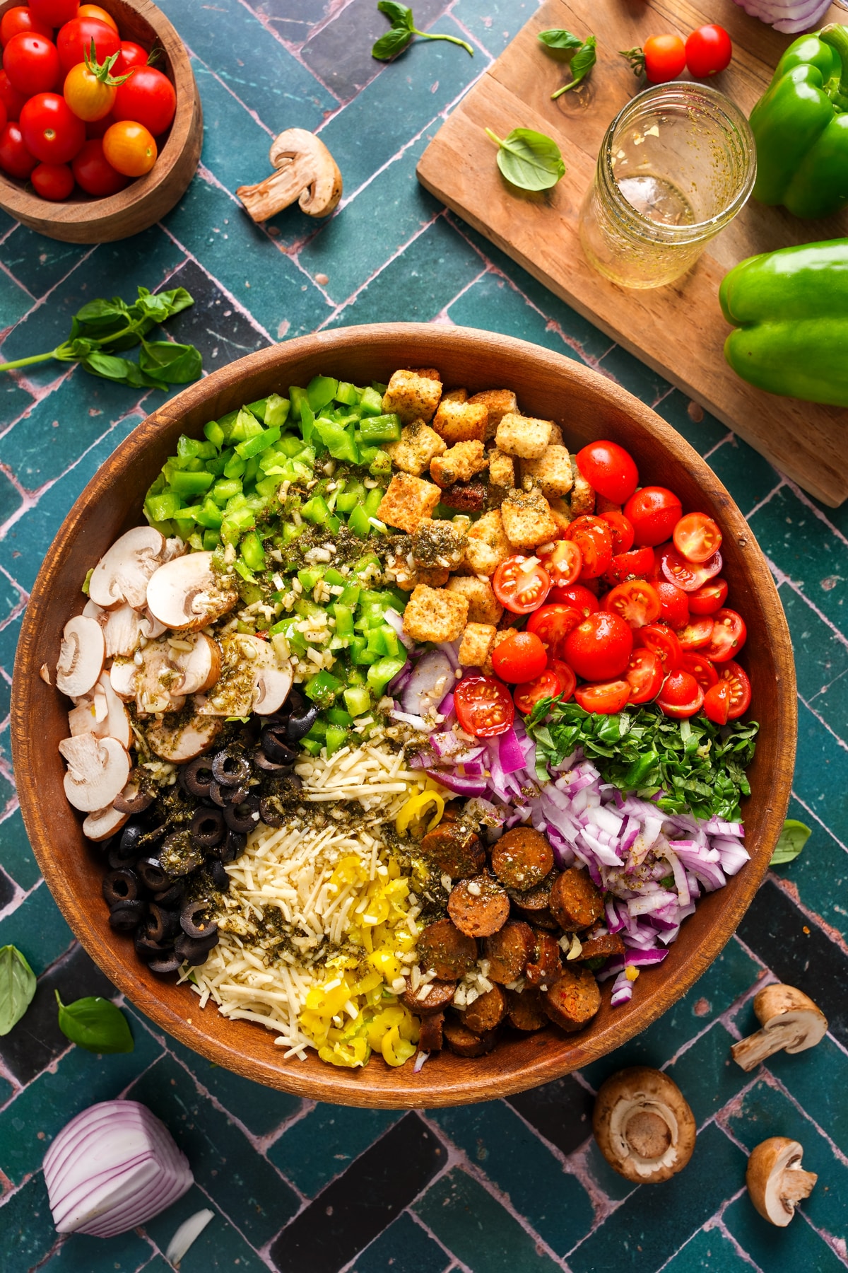 Colorful vegan pizza salad in a wooden bowl with fresh cherry tomatoes, black olives, mushrooms, green bell peppers, red onions, vegan cheese, plant-based sausage, and croutons on a green tiled surface with fresh ingredients scattered around