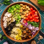 Overhead view of a deconstructed vegan pizza salad in a wooden bowl with neatly arranged rows of cherry tomatoes, croutons, green bell peppers, mushrooms, olives, vegan cheese, banana peppers, vegan sausage, red onion, and fresh basil.