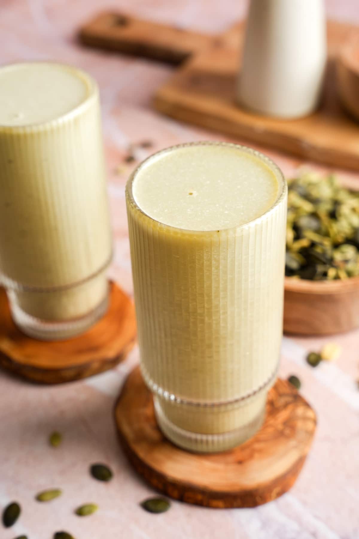 Two tall glasses filled with homemade pumpkin seed milk, placed on wooden coasters with scattered pumpkin seeds and a soft pink background.