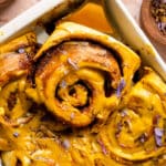 Close-up of a golden milk cinnamon roll being lifted from a baking dish, swirled with turmeric glaze and sprinkled with dried edible flower petals.