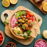 Flat lay of a vibrant Mediterranean-inspired falafel bowl on a wooden board, featuring white rice, homemade falafel, hummus, cherry tomatoes, cucumbers, pickled onions, olives, and pita bread. Surrounded by lemons, fresh parsley, and small bowls of olives and onions for garnish. Bright, clean, and styled for a healthy vegan recipe blog.