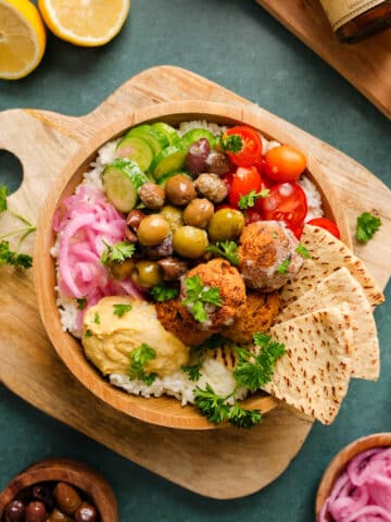 Flat lay of a vibrant Mediterranean-inspired falafel bowl on a wooden board, featuring white rice, homemade falafel, hummus, cherry tomatoes, cucumbers, pickled onions, olives, and pita bread. Surrounded by lemons, fresh parsley, and small bowls of olives and onions for garnish. Bright, clean, and styled for a healthy vegan recipe blog.