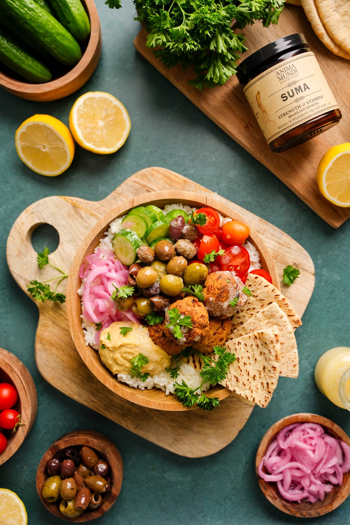Vibrant vegan falafel bowl filled with white rice, crispy falafel, creamy hummus, cherry tomatoes, cucumbers, kalamata and green olives, pickled red onions, pita slices, and fresh parsley, styled on a wooden board with lemons and Anima Mundi SUMA supplement in the background