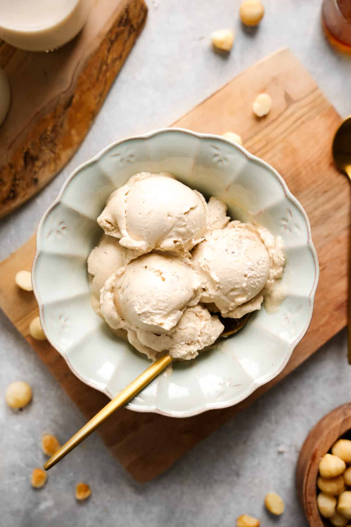 Homemade vegan macadamia nut ice cream in a scalloped ceramic bowl, served with a gold spoon. This creamy, dairy-free ice cream is made with coconut milk and macadamia butter for a rich, smooth texture. Easy no-blender recipe, perfect for summer dessert or Ninja Creami use. Gluten-free, refined sugar-free, and naturally sweetened.