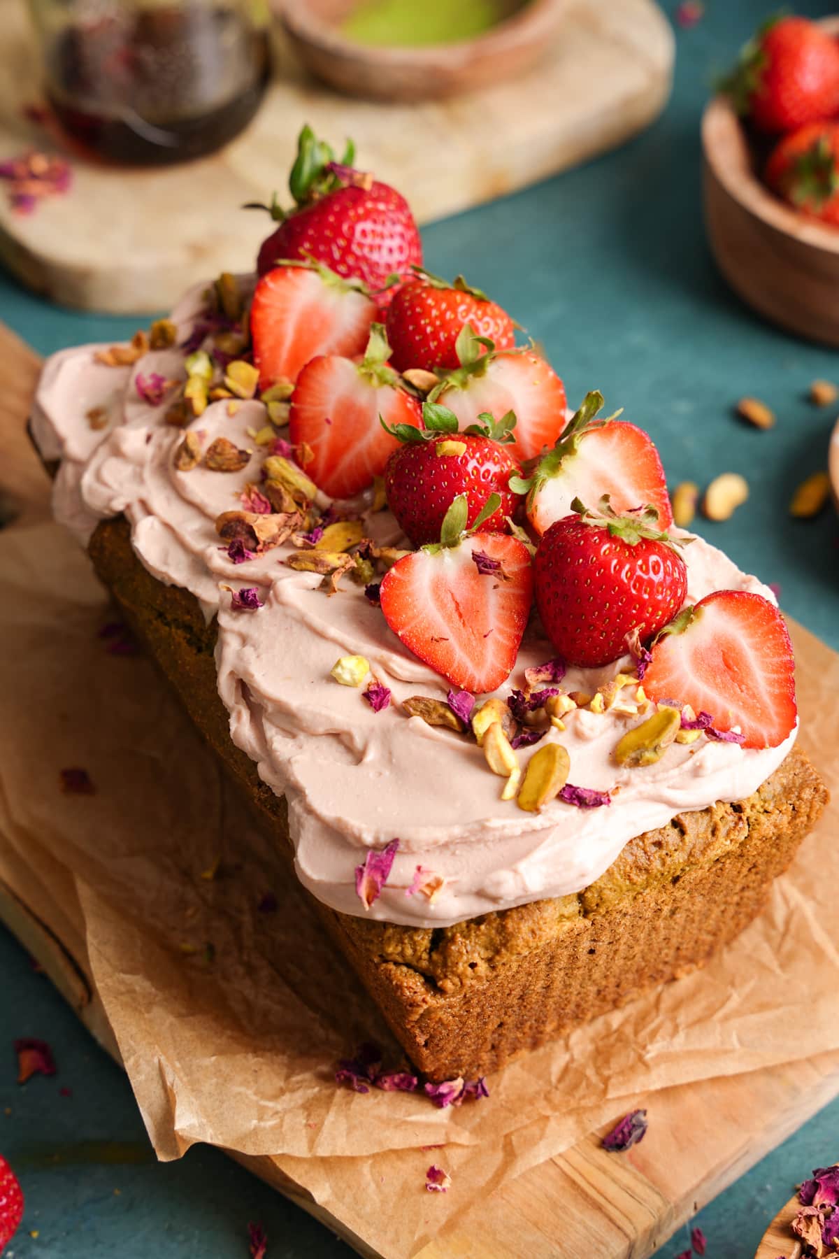 Close-up of a beautifully decorated vegan pistachio rose cake topped with fluffy rose-pink whipped frosting, halved fresh strawberries, crushed pistachios, and edible rose petals, sitting on parchment paper atop a wooden board.