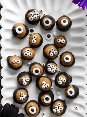 Vegan SunButter chocolate eyeballs arranged on a white scalloped platter, each coated in dark chocolate with candy eyes on top for a fun Halloween treat, surrounded by black lace and colorful flowers.