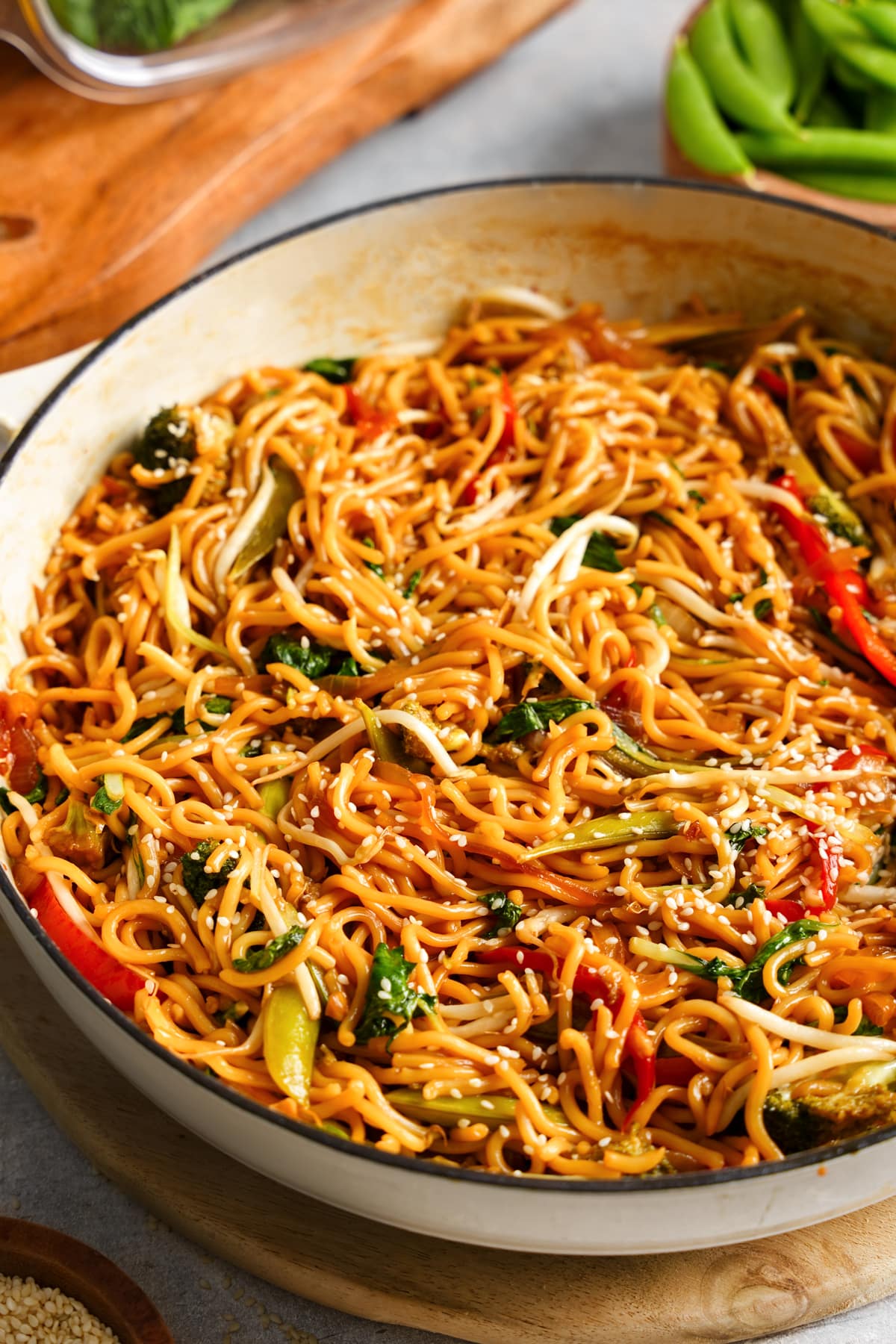 **Alt Text:**
A close-up of vegan chow mein in a large white pot, featuring stir-fried noodles tossed with colorful vegetables like bell peppers, bok choy, broccoli, and bean sprouts. The dish is coated in a glossy soy-based sauce and sprinkled with sesame seeds, highlighting its vibrant and savory presentation.