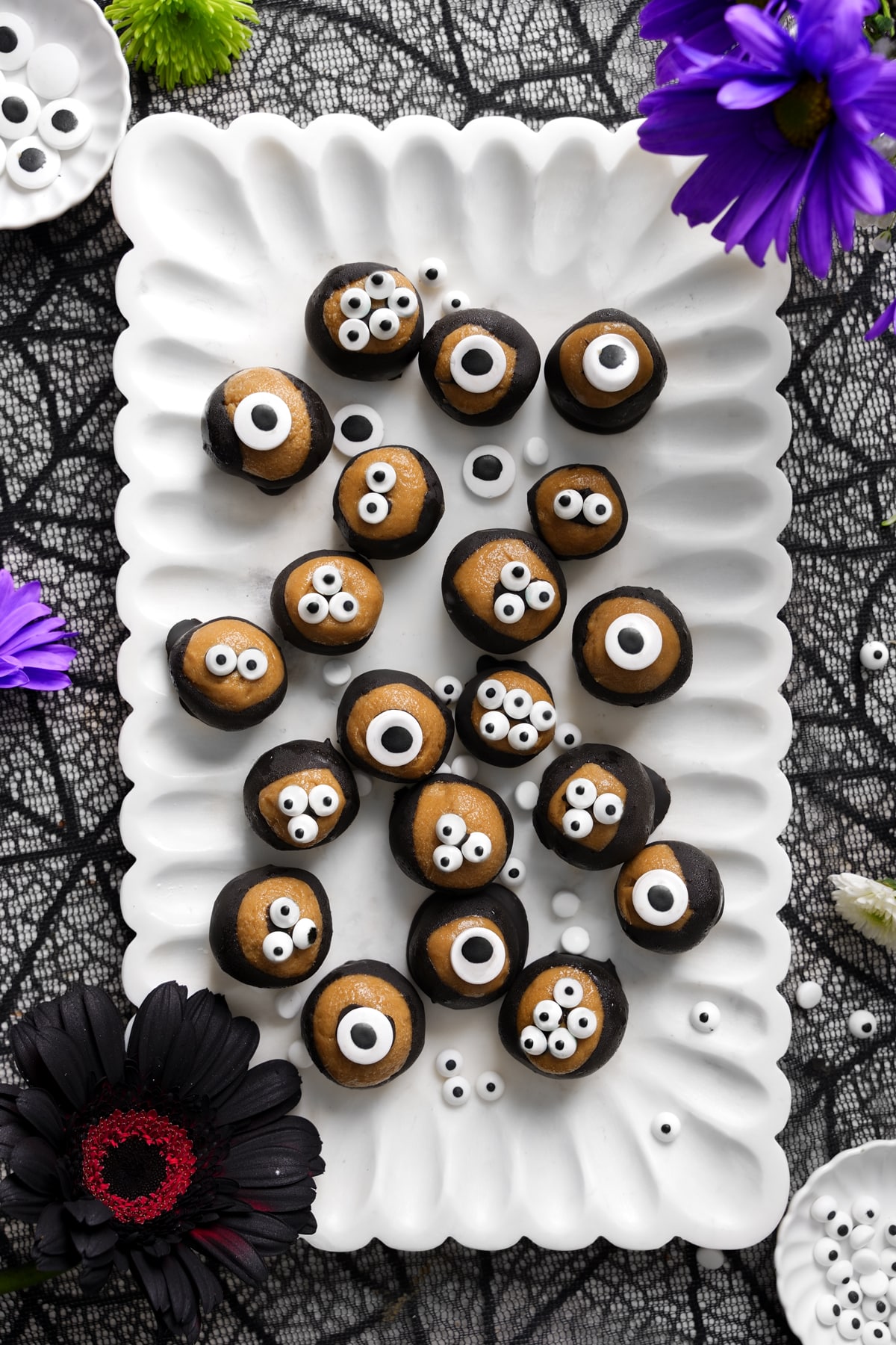 Vegan SunButter chocolate eyeballs arranged on a white scalloped platter, each coated in dark chocolate with candy eyes on top for a fun Halloween treat, surrounded by black lace and colorful flowers.