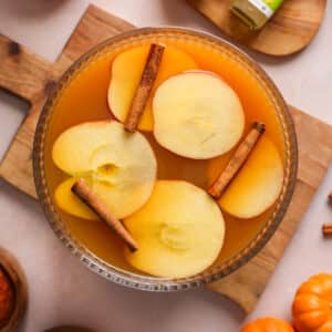 Overhead view of a glass punch bowl filled with spiced apple pumpkin cider topped with apple slices and cinnamon sticks, styled on a wooden board with autumn decor