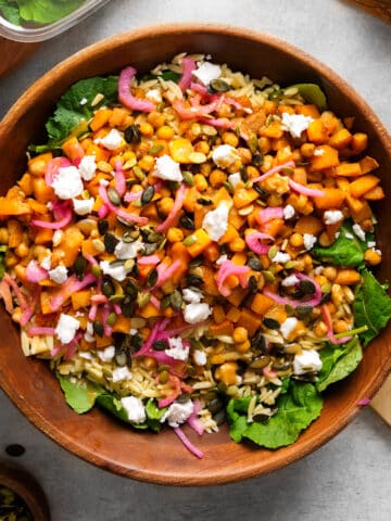A vibrant vegan butternut squash and kale salad served in a wooden bowl, featuring roasted squash, chickpeas, orzo, pickled red onions, pumpkin seeds, and crumbled vegan feta over fresh baby kale. The colorful mix of ingredients creates a nourishing, hearty fall-inspired salad.