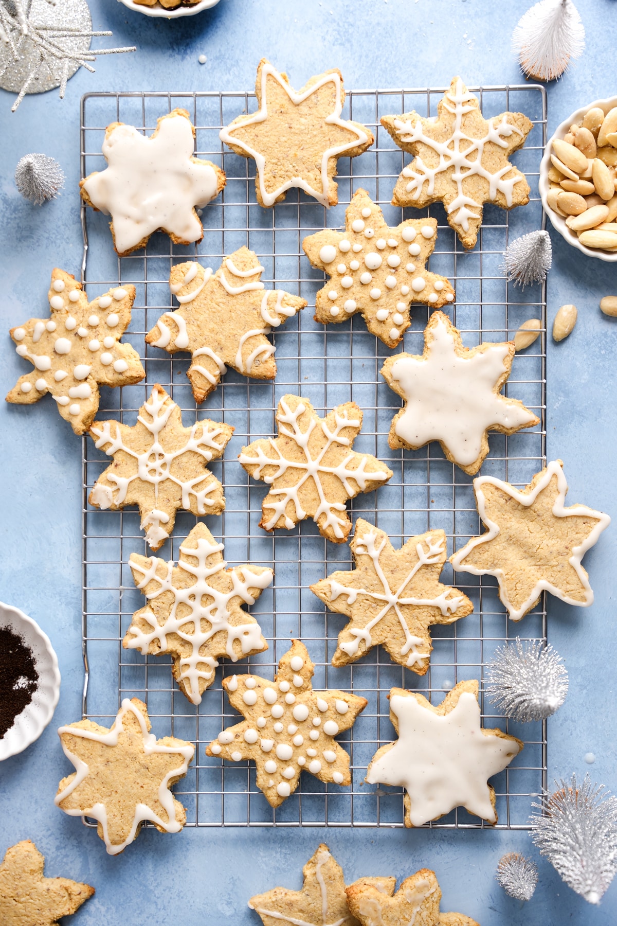 A cooling rack filled with almond flour sugar cookies shaped like snowflakes, decorated with white icing in various patterns such as dots, lines, and intricate snowflake designs. The cookies sit on a light blue surface with small holiday decorations, including silver bottle-brush trees and bowls of almonds scattered around.