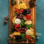 Christmas tree–shaped charcuterie board with tater tots, cherry tomatoes, grapes, olives, cucumbers, berries, nuts, cheese slices, hummus, and dips arranged with fresh rosemary on a wooden board.
