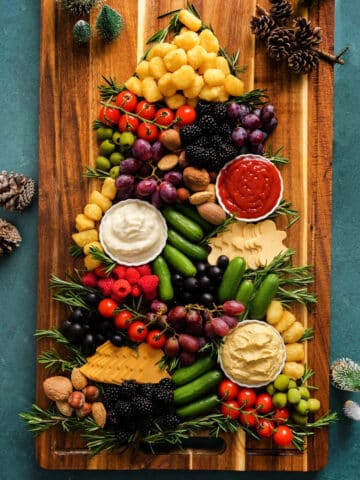 Christmas tree–shaped charcuterie board with tater tots, cherry tomatoes, grapes, olives, cucumbers, berries, nuts, cheese slices, hummus, and dips arranged with fresh rosemary on a wooden board.
