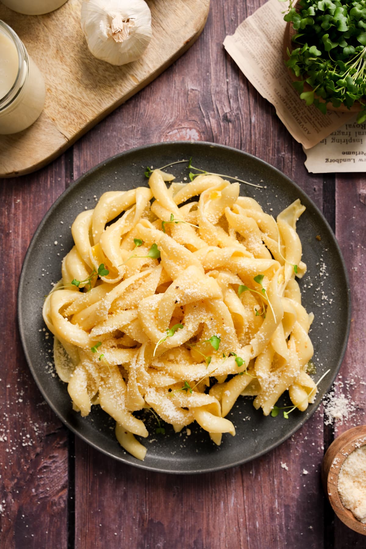 A dark plate filled with creamy truffle pasta topped with vegan parmesan and microgreens, set on a rustic wooden surface with garlic, grated cheese, and fresh microgreens surrounding the dish.