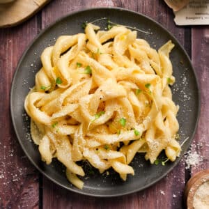 A top view of a plate of creamy vegan truffle pasta topped with dairy-free parmesan and microgreens, served on a dark plate over a rustic wooden table.