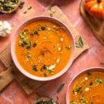 Overhead view of creamy vegan pumpkin soup in a pink bowl, garnished with pumpkin seeds, microgreens, coconut cream drizzle, and black pepper, set on a wooden board with garlic, sage leaves, and small pumpkins on a warm terracotta background.