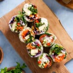 Fresh vegan tofu spring rolls sliced in half and arranged on a wooden cutting board, filled with rice noodles, tofu, shredded carrots, red cabbage, cucumber, microgreens, and herbs on a light blue background.