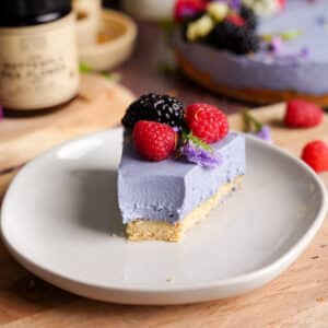 Close-up of a vegan butterfly pea flower cheesecake slice with a smooth purple filling and shortbread crust, topped with raspberries, blackberry, and edible flowers on a neutral plate.