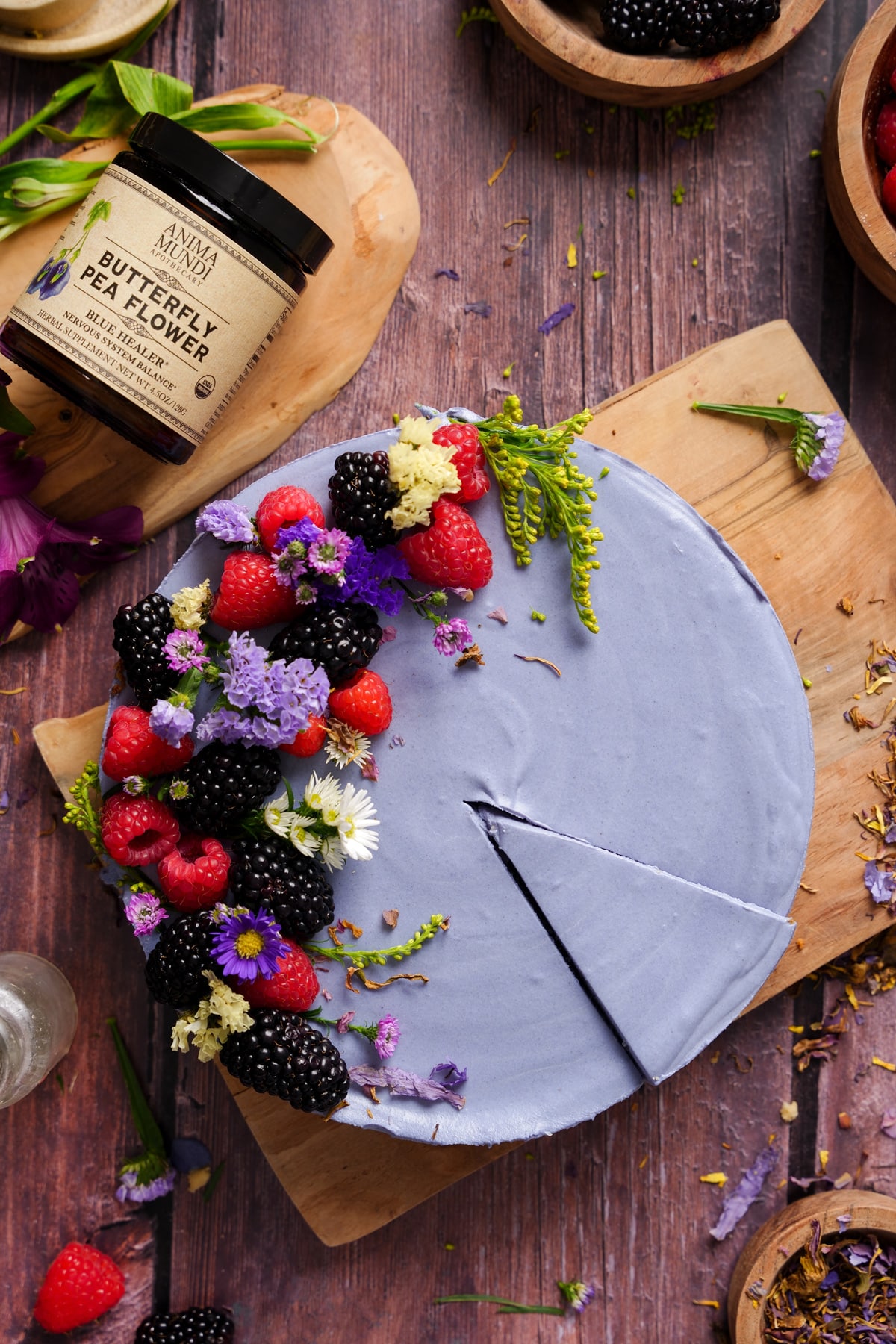 Blue butterfly pea flower cheesecake with natural purple coloring, topped with fresh raspberries, blackberries, and edible flowers on a wooden board - a vibrant vegan floral dessert made using butterfly pea powder