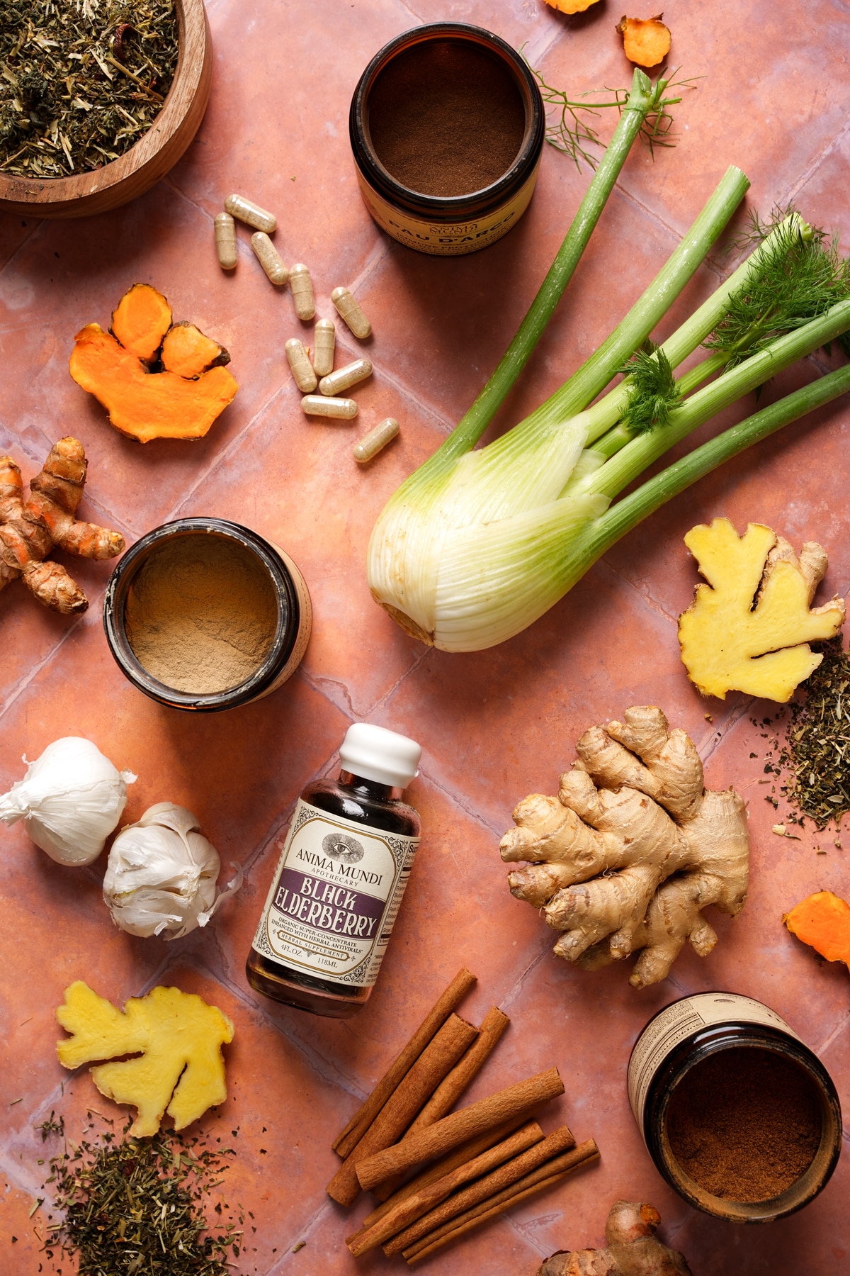 Flat lay of immune-supporting herbs and ingredients including fennel bulb, fresh ginger root, turmeric, garlic cloves, cinnamon sticks, dried herbs, supplement capsules, and an Anima Mundi Black Elderberry bottle arranged on a warm terracotta surface.