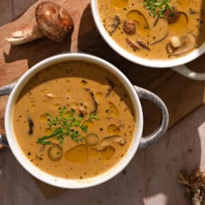 Two bowls of creamy vegan mushroom soup topped with fresh chives and olive oil, served with whole mushrooms on a wooden board - easy dairy-free mushroom soup recipe.