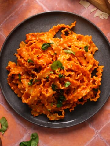 Overhead view of mafaldine pasta coated in a creamy dairy-free vodka sauce, garnished with fresh basil and served in a dark bowl on a warm terracotta surface.