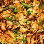 Close-up of saucy baked dumplings topped with chopped peanuts, sesame seeds, chili oil, wilted greens, and sliced green onions. The dumplings are golden around the edges and coated in a rich savory sauce, creating a vibrant Asian-inspired dumpling bake.
