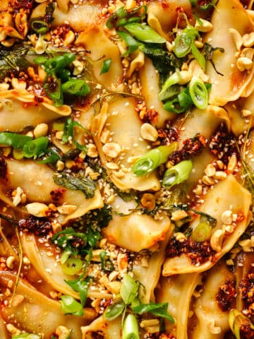 Close-up of saucy baked dumplings topped with chopped peanuts, sesame seeds, chili oil, wilted greens, and sliced green onions. The dumplings are golden around the edges and coated in a rich savory sauce, creating a vibrant Asian-inspired dumpling bake.