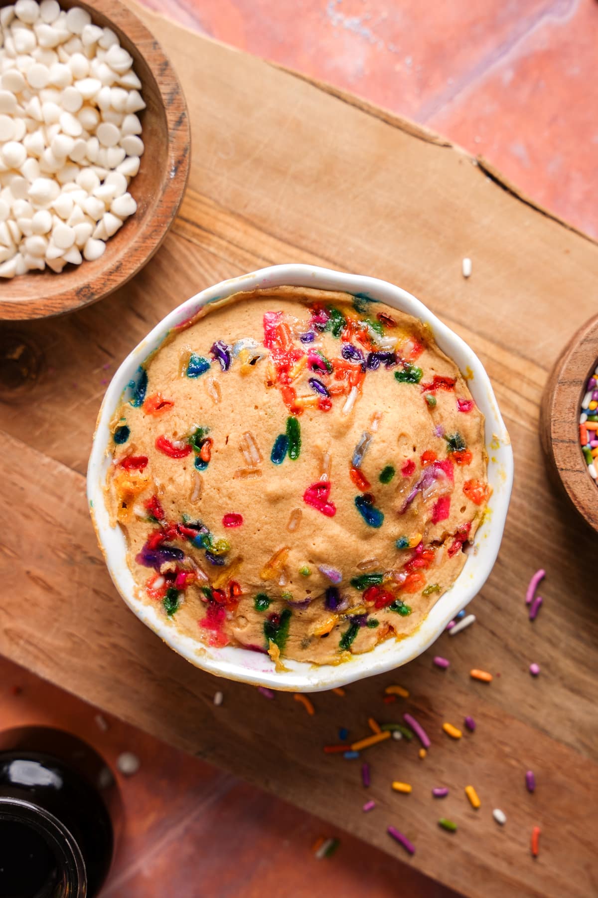 High-protein vanilla mug cake with colorful sprinkles in a white mug on a wooden board, surrounded by white chocolate chips and baking ingredients.