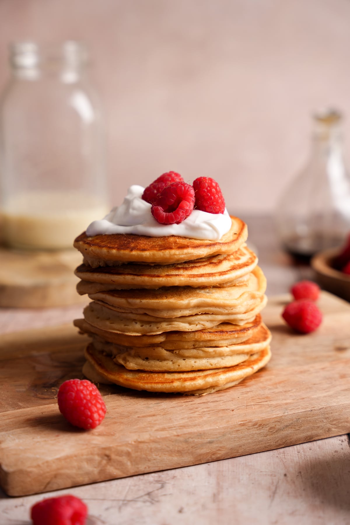 Stack of fluffy golden pancakes topped with a dollop of whipped cream and fresh raspberries, served on a wooden board with a soft, neutral background.