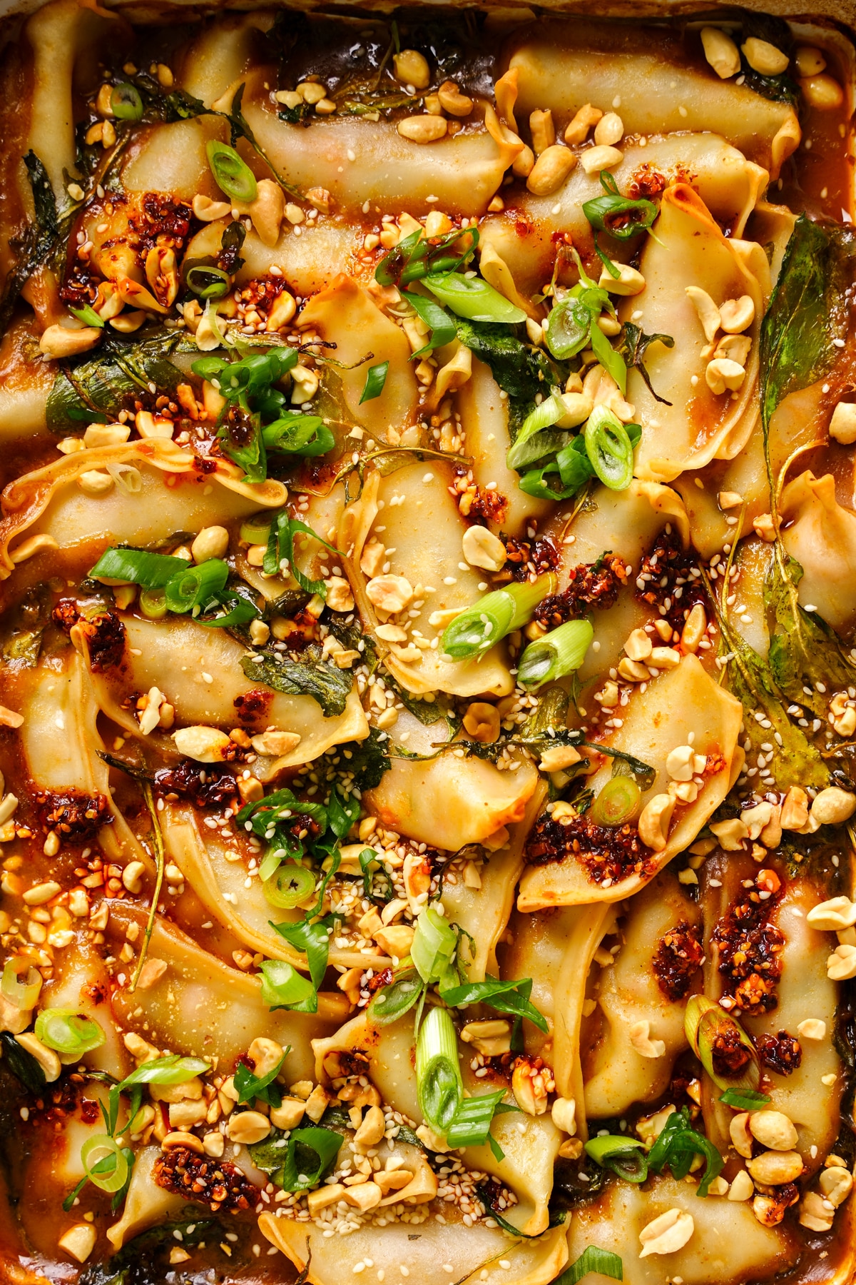 Overhead close-up of a baked dumpling dish topped with chopped peanuts, sliced green onions, sesame seeds, chili oil, and wilted greens in a rich savory sauce. The golden dumplings are slightly crisp on the edges and arranged tightly in a baking dish, creating a colorful, textured Asian-inspired dumpling bake.