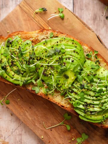 Toasted slice of rustic bread topped with sliced avocado, microgreens, and sprinkled seasoning, served on a wooden board.