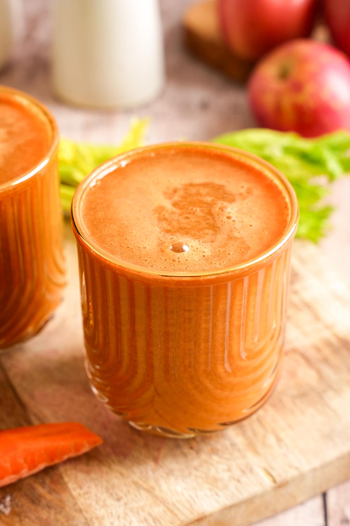 Fresh carrot celery juice served in a glass with a frothy top, surrounded by whole carrots, apples, celery, and a bottle of juice in the background.