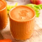 Fresh carrot celery juice served in a glass with a frothy top, surrounded by whole carrots, apples, celery, and a bottle of juice in the background.