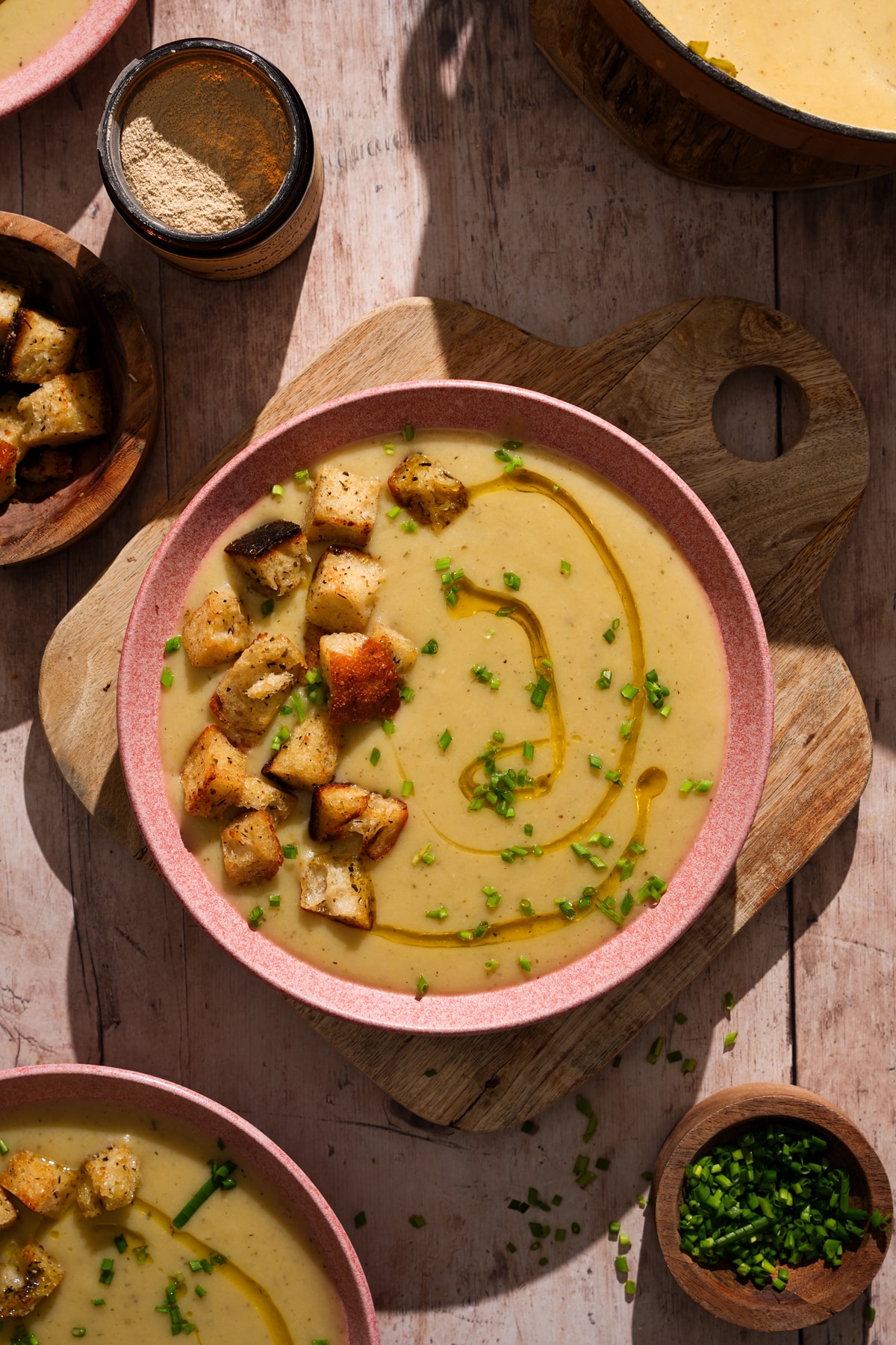 Creamy vegan potato leek soup topped with crispy croutons, fresh chives, and olive oil with ingredients and seasonings on a rustic table