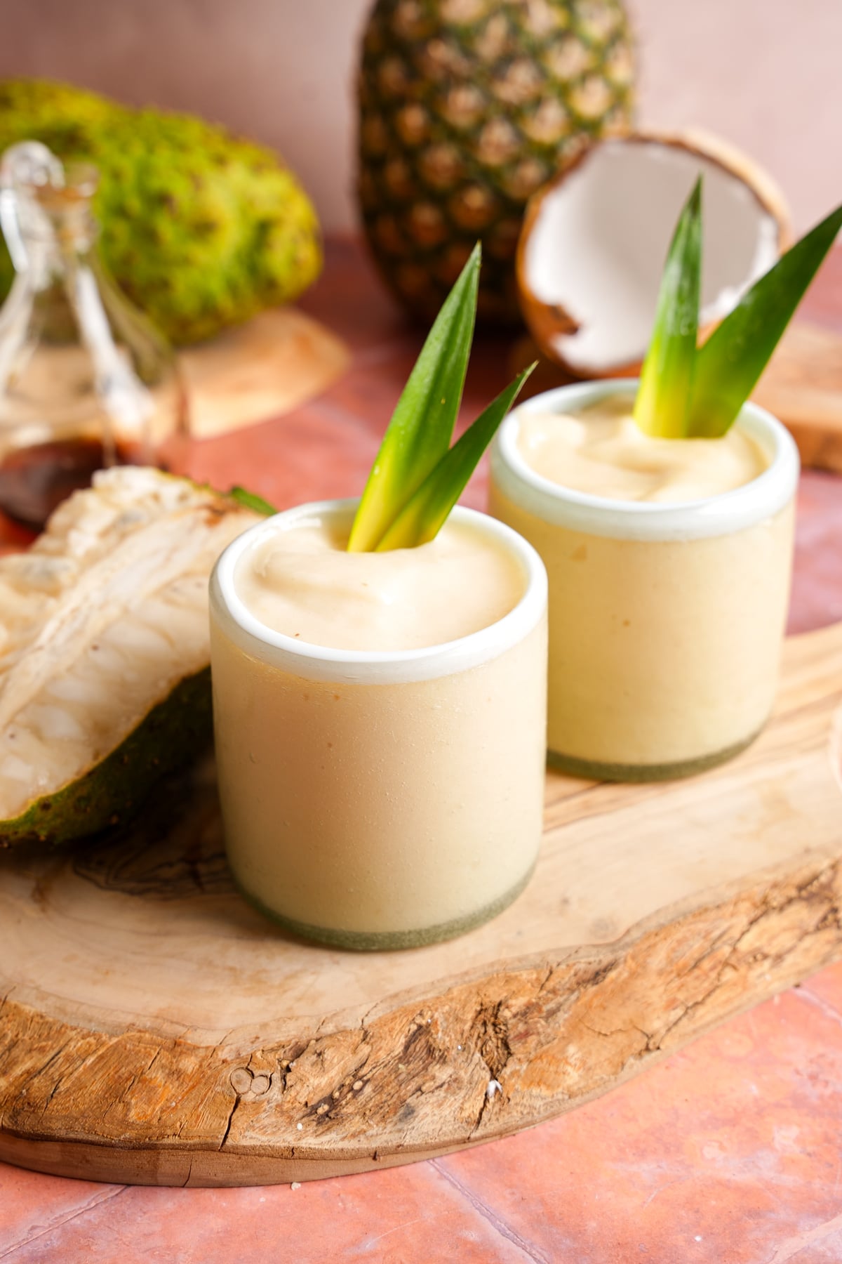 Two creamy soursop smoothies served in small glasses, garnished with fresh pineapple leaves and set on a rustic wooden board with tropical fruits in the background.
