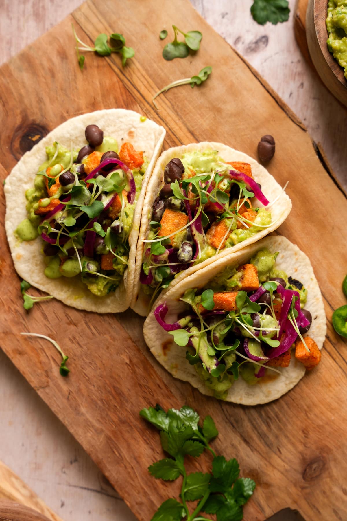 Three soft tortillas filled with seasoned sweet potato croutons, black beans, guacamole, pickled red onions, microgreens, and a creamy green cilantro lime dressing, served on a rustic wooden board with fresh cilantro scattered around.