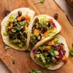 Three open-faced sweet potato tacos on small tortillas topped with guacamole, black beans, pickled red onions, microgreens, and creamy green dressing, arranged on a wooden cutting board with a bag of sweet potato croutons blurred in the background.