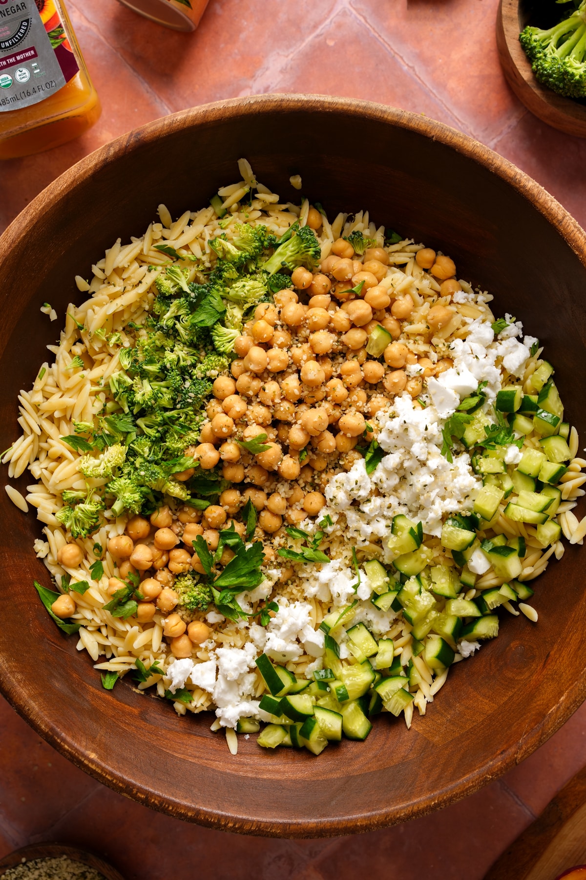 Overhead view of a wooden bowl filled with vegan orzo pasta salad, featuring cooked orzo, chickpeas, chopped broccoli, diced cucumbers, crumbled vegan feta, fresh parsley, and hemp seeds, with a light dressing visible and ingredients arranged in sections before mixing.