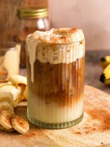 Healthy iced vegan banana latte topped with fluffy banana cold foam and a sprinkle of cinnamon. This creamy dairy-free coffee drink is layered with espresso and plant-based milk, served with fresh banana slices for an easy homemade café-style beverage.