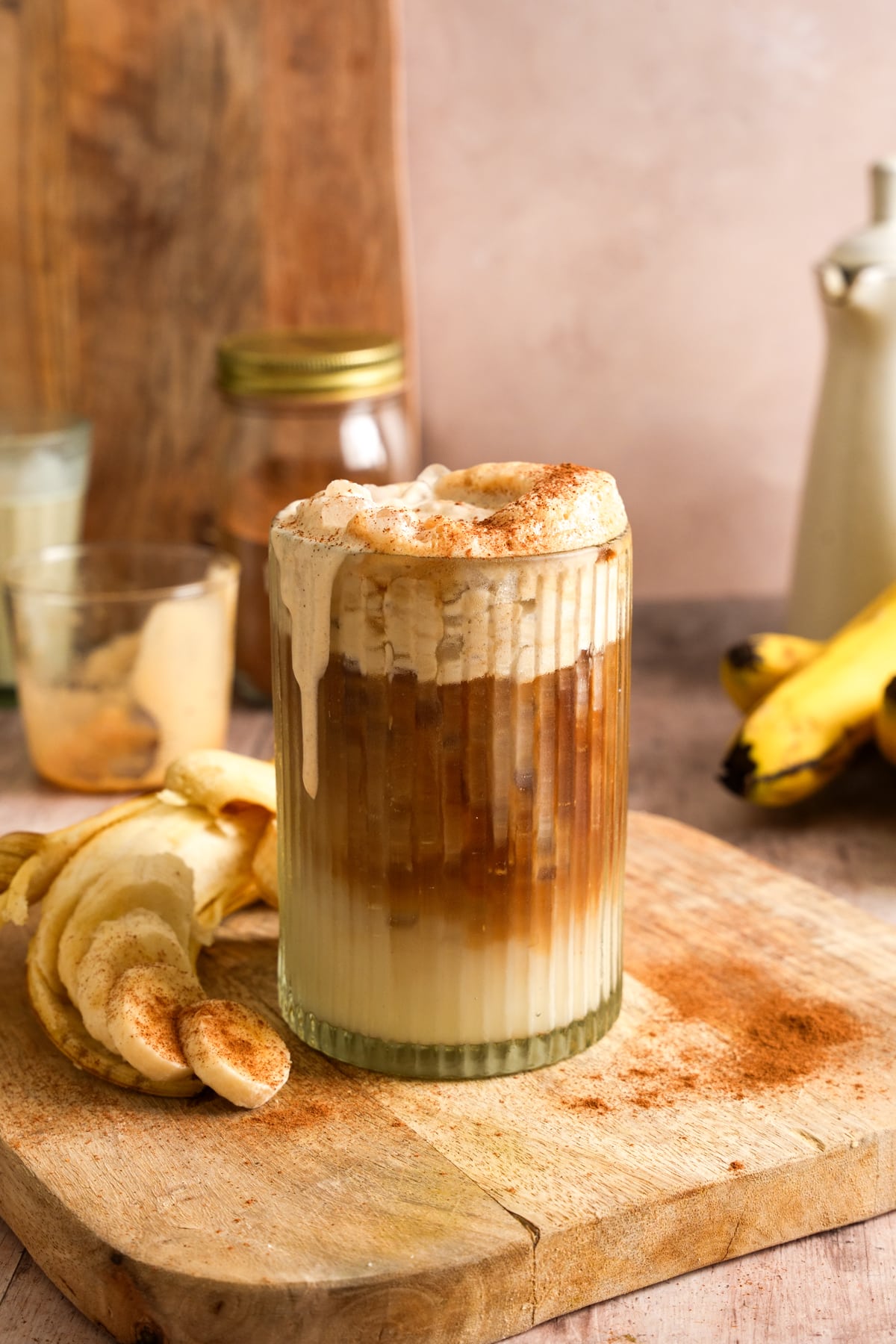 Glass of iced banana coffee topped with creamy banana cold foam and a dusting of cinnamon, served on a wooden board with fresh banana slices and a peeled banana. This healthy homemade banana coffee recipe features layers of espresso and creamy plant-based milk, making it a refreshing vegan coffee drink for breakfast or an afternoon pick-me-up.
