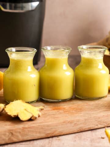 Healthy homemade ginger shots made with fresh ginger, pineapple, and lemon juice in three small glass bottles. Surrounded by fresh ginger root, lemons, and a juicer, these immune-boosting wellness shots are perfect for meal prep and daily nutrition.