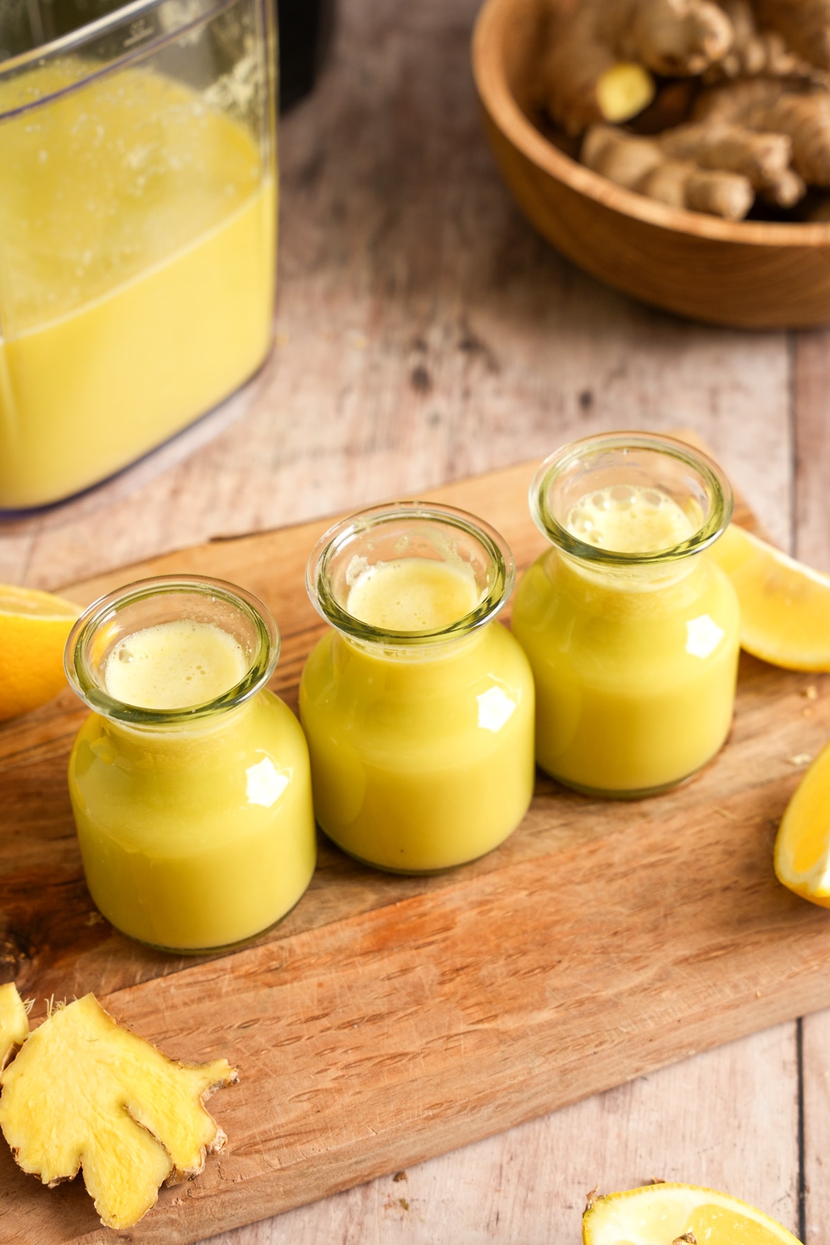 Homemade ginger wellness shots made with fresh ginger, pineapple, and lemon juice poured into small glass bottles on a wooden board. Fresh ginger root, pineapple slices, lemons, and a blender of juice surround these healthy immune-boosting ginger shots.