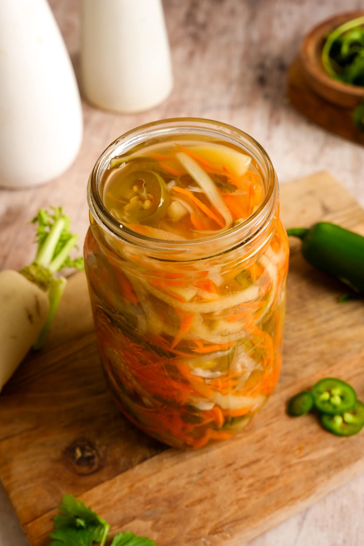 A glass jar filled with homemade pickled vegetables sits on a wooden cutting board. The colorful mix includes shredded carrots, sliced onions, jalapeño peppers, and other vegetables submerged in a clear pickling brine. Fresh jalapeño slices, herbs, and root vegetables are arranged around the jar, creating a rustic kitchen scene.