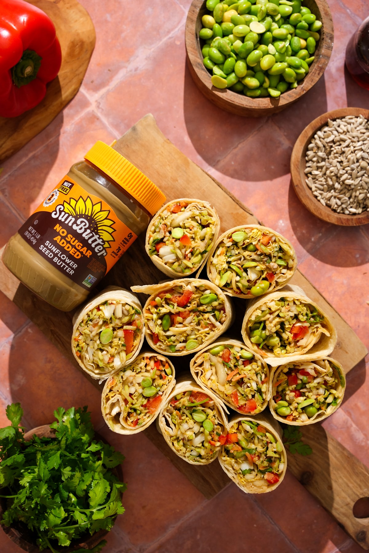 Overhead view of sliced vegan Asian crunch wraps arranged on a wooden serving board. The wraps are filled with Napa cabbage, broccoli slaw, edamame, red bell pepper, cilantro, and a creamy sunflower seed butter dressing. A jar of SunButter No Sugar Added Sunflower Seed Butter sits beside the wraps, with bowls of edamame and sunflower seeds, fresh cilantro, and a red bell pepper surrounding the board. The colorful wraps make a vibrant, plant-based lunch or meal prep option.