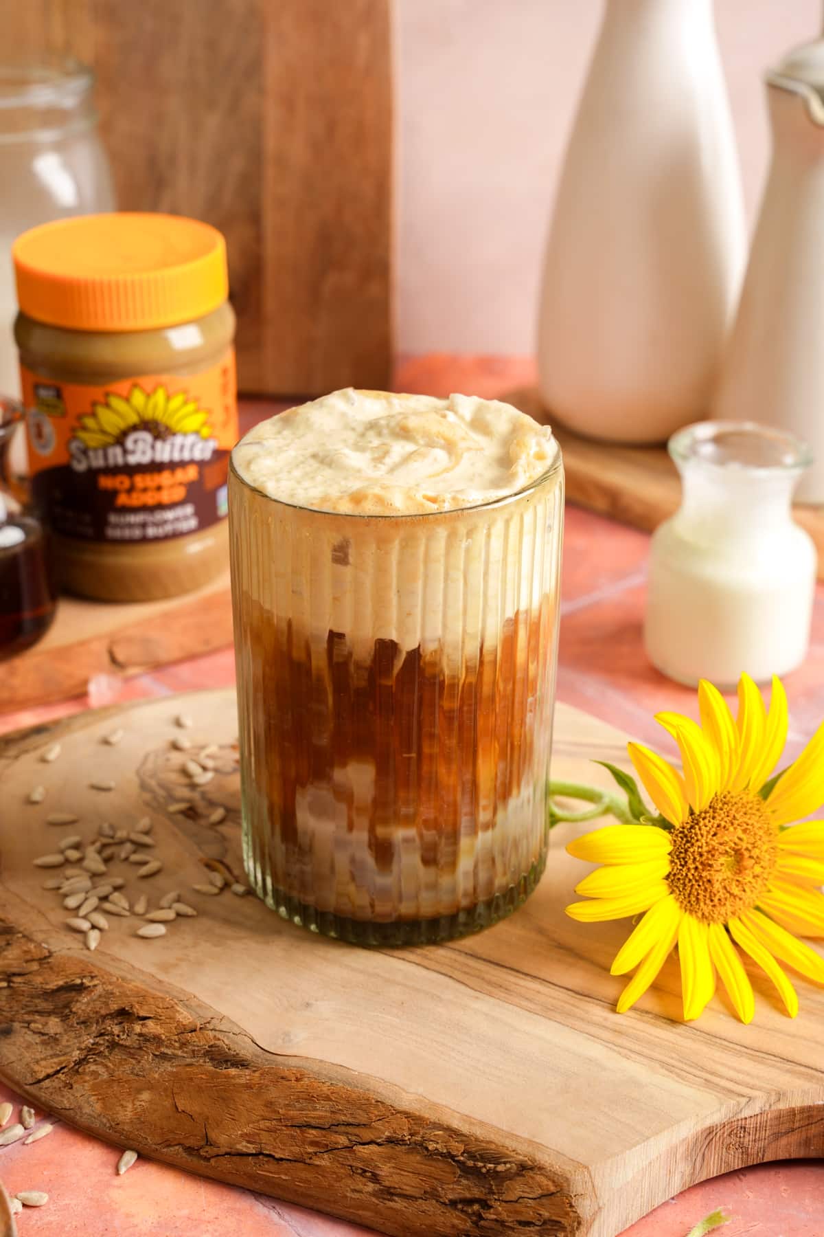 A creamy iced SunButter latte served in a ribbed glass with layers of espresso, dairy-free milk, and fluffy SunButter cold foam. The coffee is photographed on a rustic wooden board with a jar of SunButter, sunflower seeds, maple syrup, plant-based milk, and a bright sunflower in the background.
