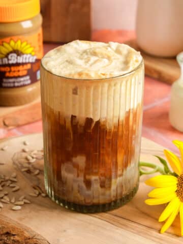 Creamy iced SunButter latte topped with thick whipped foam in a ribbed glass, served on a rustic wooden board. A jar of SunButter, plant-based milk, sunflower seeds, and a bright sunflower surround this dairy-free coffee drink for a cozy café-style presentation.